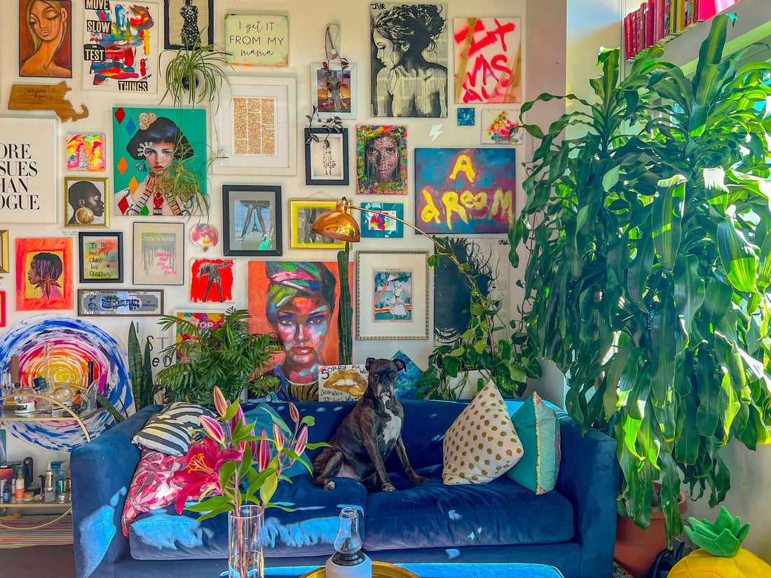 A vibrant and eclectic gallery wall in a bohemian living room featuring colorful artwork, framed prints, and paintings. A blue velvet sofa with throw pillows, lush green plants, and a black dog add charm to the artistic, cosy interior.